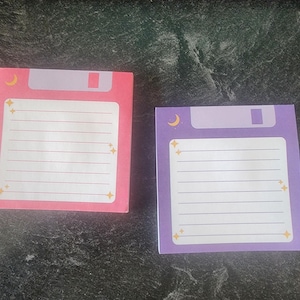May include: Two square sticky note pads designed to resemble vintage floppy disks. One pad is pink and the other is purple. Both feature a white lined writing area, a light purple top section, a small yellow crescent moon icon, and yellow star accents.