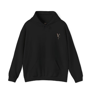 May include: A black long-sleeve hooded sweatshirt featuring a front kangaroo pocket and a drawstring hood. On the upper left chest, there is a small, vertically oriented logo in a light tan colour consisting of the letters VC with a star icon.