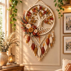 May include: A circular macrame wall hanging featuring a central tree of life design with woven branches and leaves in shades of orange, yellow, and cream. The piece is adorned with fabric flowers and hanging leaf-shaped tassels in autumn colors.