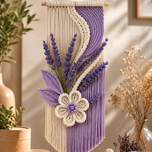 May include: A macrame wall hanging featuring a split design of cream and purple vertical cords. The piece is adorned with a central cream-colored flower with a purple center, two purple macrame leaves, and several sprigs of purple lavender.