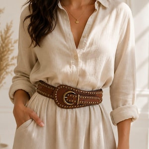 May include: A beige long-sleeved button-down shirt dress with a collar and pockets. The dress is cinched at the waist with a wide brown leather belt featuring small metallic studs and a large brass-toned buckle with a central prong closure.