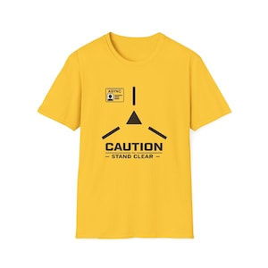 May include: A bright yellow short-sleeved t-shirt featuring a black graphic print. The design includes a small rectangular ID badge labeled ASYNC, a central black triangle surrounded by three radiating lines, and the text CAUTION STAND CLEAR.
