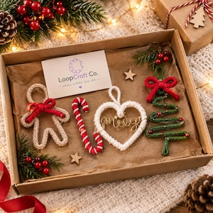 Puede incluir: Una caja de regalo de cartón que contiene cuatro adornos navideños de ganchillo: un hombre de jengibre beige con un lazo rojo, un bastón de caramelo rojo y blanco, un corazón blanco con texto en hilo dorado y un árbol verde con cuentas rojas.