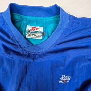 May include: A royal blue pullover windbreaker with a ribbed collar and a teal interior lining. The inner label reads Headwind Sun Mountain Sports with a flag graphic. A small white and brown tree embroidery is visible on the lower right chest area.