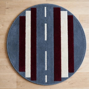 May include: A circular rug featuring a blue background with a central vertical white dashed line resembling a road. The design includes symmetrical vertical stripes in deep burgundy and off-white, creating a structured geometric pattern across the surface.