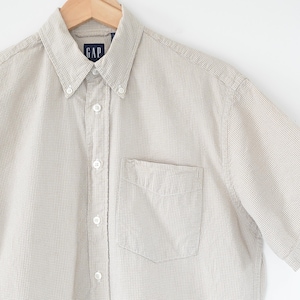 Può includere: Camicia button-down a maniche corte con motivo a quadri beige chiaro e bianco. La camicia ha un colletto button-down, una tasca sul petto e il logo "GAP" sul colletto. La camicia è appesa a una gruccia di legno.