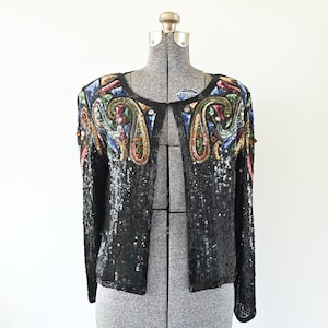 May include: Black sequined jacket with colorful paisley and floral embroidery on the shoulders and upper chest. The long-sleeved jacket is open in the front and displayed on a mannequin.