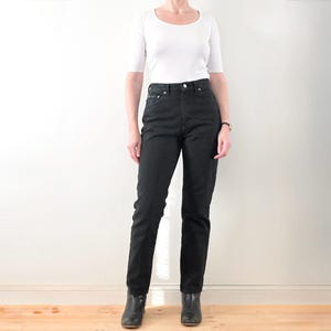 Peut inclure: Une personne porte un haut blanc à col rond et des jeans noirs. Les jeans sont taille haute et droits. La personne porte également des bottines noires.