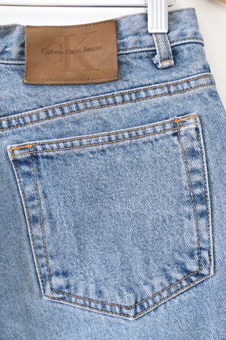 Może przedstawiać: Jasne niebieskie dżinsy z brązową sk&oacute;rzaną naszywką z napisem "Calvin Klein Jeans". Dżinsy mają tylną kieszeń z brązowym szwem.