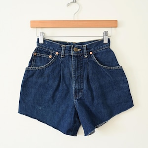 Pode incluir: Calções de ganga azul escuro com cintura alta e bainha crua. Os calções têm bolsos frontais e estão pendurados num cabide de madeira. Os calções são um estilo clássico.