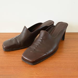 May include: A pair of brown leather mules with square toes and small heels. The shoes have a slip-on design and decorative stitching on the vamp. The shoes are displayed on a wooden surface.