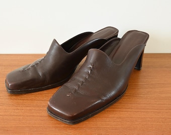 Puede incluir: Un par de mulas de cuero marrón con punteras cuadradas y tacones pequeños. Los zapatos tienen un diseño sin cordones y costuras decorativas en la pala. Los zapatos se exhiben sobre una superficie de madera.