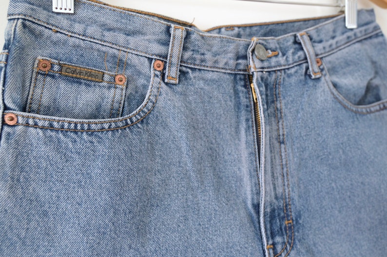 Może przedstawiać: Jasne niebieskie dżinsy z zamkiem błyskawicznym i metką Calvin Klein na przedniej kieszeni.