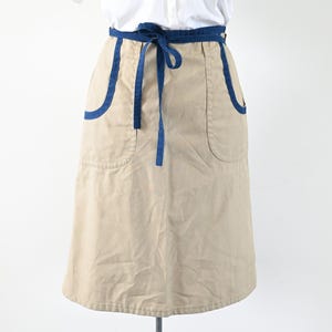 Könnte beinhalten: Beige Schürze mit marineblauen Details. Die Schürze hat zwei große Vordertaschen und marineblaue Paspeln. Sie hat eine marineblaue Bindeband. Die Schürze ist aus robustem Stoff gefertigt, geeignet für Küche oder Bastelarbeiten.