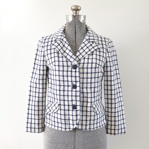 Puede incluir: Una blazer a cuadros blancos y azul marino con cuello con solapas, tres botones azul marino y dos bolsillos delanteros. La chaqueta se exhibe en un maniquí.