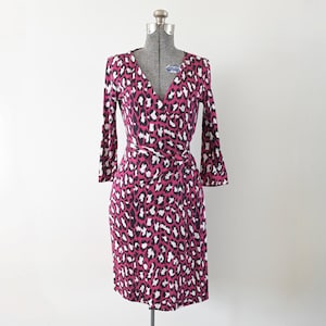 May include: A wrap dress with a V-neck and long sleeves. The dress features a pink, black, and white animal print. The dress is displayed on a mannequin against a white background. The dress is knee-length.