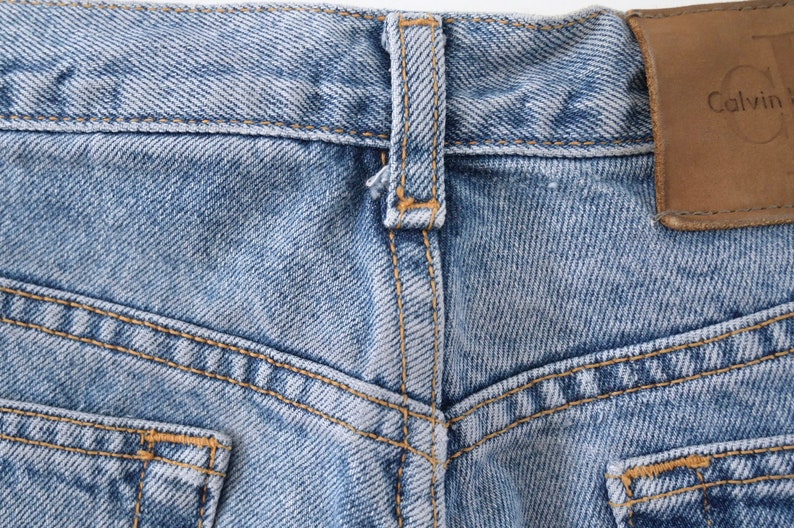 Może przedstawiać: Zbliżenie na parę jasnoniebieskich dżins&oacute;w z brązową sk&oacute;rzaną naszywką z napisem "Calvin Klein" na tylnym pasie.