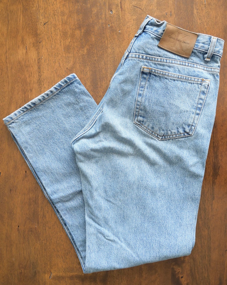 Może przedstawiać: Jasne niebieskie dżinsy z brązową sk&oacute;rzaną naszywką z napisem "Calvin Klein Jeans" na tylnej kieszeni.
