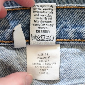Może przedstawiać: Biała etykieta pielęgnacyjna z czarnym tekstem i symbolami. Tekst brzmi "Wash separately before wearing. Designed to fade and lose color. Turn inside out. Machine wash warm. Can be dry cleaned. RN 36009". Pod tekstem znajduje się symbol prania w pralce, symbol wybielania, symbol suszenia w suszarce bębnowej i symbol prasowania. Druga biała etykieta z czarnym tekstem brzmi "Size: 13, Inseam: 32, BJXJ137, Straight Leg Jean, Double Stone, 33897, 010158".
