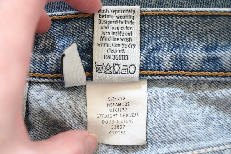 Może przedstawiać: Biała etykieta pielęgnacyjna z czarnym tekstem i symbolami. Tekst brzmi "Wash separately before wearing. Designed to fade and lose color. Turn inside out. Machine wash warm. Can be dry cleaned. RN 36009". Pod tekstem znajduje się symbol prania w pralce, symbol wybielania, symbol suszenia w suszarce bębnowej i symbol prasowania. Druga biała etykieta z czarnym tekstem brzmi "Size: 13, Inseam: 32, BJXJ137, Straight Leg Jean, Double Stone, 33897, 010158".