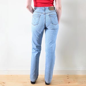 Könnte beinhalten: Hellblaue Jeans mit klassischem geradem Bein. Die Jeans haben das Calvin Klein-Logo auf den Gesäßtaschen und einen Lederaufnäher am Bund. Das Model trägt ein rotes Oberteil.