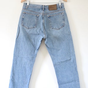 Może przedstawiać: Jasne niebieskie dżinsy wiszące na drewnianym wieszaku. Dżinsy mają brązową sk&oacute;rzaną naszywkę z tyłu z napisem "Calvin Klein Jeans".