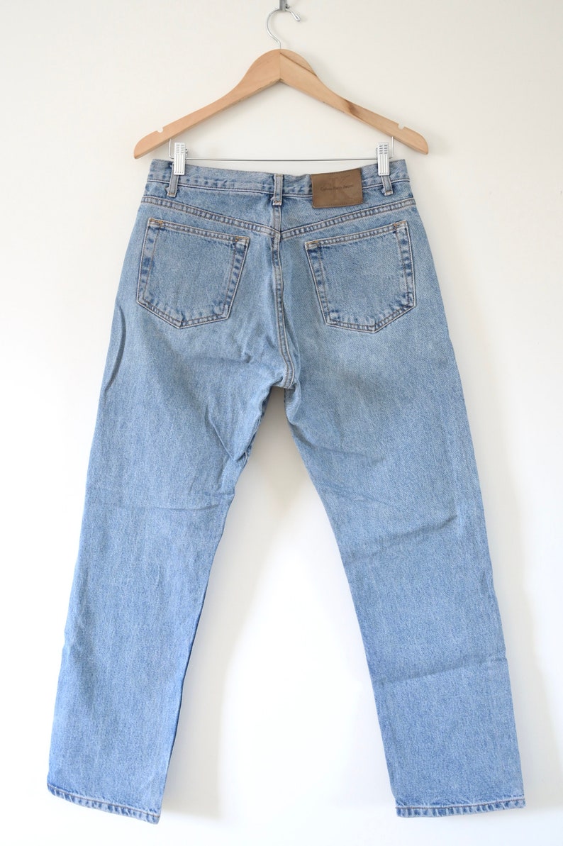 Może przedstawiać: Jasne niebieskie dżinsy wiszące na drewnianym wieszaku. Dżinsy mają brązową sk&oacute;rzaną naszywkę z tyłu z napisem "Calvin Klein Jeans".