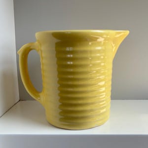 May include: A bright yellow ceramic pitcher featuring a cylindrical body with a distinct horizontal ribbed texture, a curved handle on the left side, and a small pouring spout on the right side, finished with a smooth, glossy glaze.