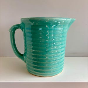 May include: A turquoise ceramic jug with a glossy finish and a ribbed, horizontal texture along the body. The vessel features a curved handle on the left side and a small pouring spout on the right side, showcasing a consistent colour.
