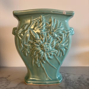 Könnte beinhalten: Eine helltürkise Keramikvase mit glänzender Oberfläche, die auf der Vorderseite ein erhabenes Reliefdesign aus Chrysanthemenblüten aufweist. Die Vase hat einen ausgestellten Rand, einen konischen Körper und kleine Seitengriffe mit einem weißen Abplatzer.