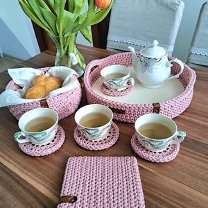 May include: A set of handmade pink crocheted home accessories including a round tray with a white base, a matching basket containing bread, three circular coasters, and a square mat. Each item features a small brown leather accent tag.