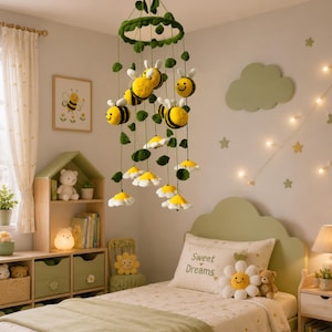 Op de afbeelding: Een hangende mobiel met gehaakte gele en zwarte bijen en witte bloemen met gele harten, opgehangen aan een groen cirkelvormig frame versierd met groene blaadjes. De mobiel hangt aan een groen koord en bevat meerdere bungelende elementen.