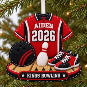 May include: A felt bowling-themed Christmas ornament featuring a red jersey with the name AIDEN and the year 2026 in white text. It includes a black bowling ball, a white pin, and red and black bowling shoes on a lane base labeled KINGS BOWLING.