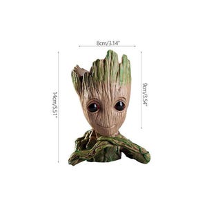 May include: A tree-like character planter with a textured brown and green bark finish, large dark eyes, and a small smile. The planter measures 8 cm (3.14 inches) in width, 9 cm (3.54 inches) in depth, and 14 cm (5.51 inches) in total height.