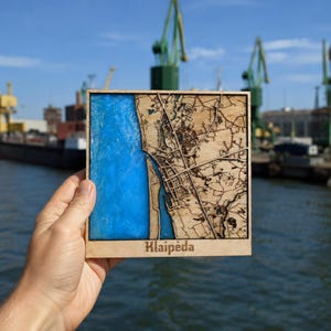 Op de afbeelding: Een vierkante houten kaart van Klaipėda met een lasergesneden stratenplan aan de rechterkant en een gestructureerd, felblauw harsgedeelte dat het water aan de linkerkant voorstelt. De naam Klaipėda is onderaan in het midden gegraveerd.