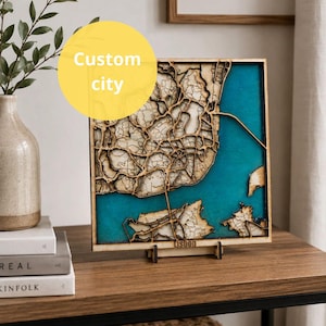 Puede incluir: Un mapa cuadrado de madera de Lisboa con patrones de calles cortados con láser sobre un fondo de agua azul turquesa vibrante. El mapa está montado sobre un pequeño soporte de madera e incluye el texto Lisbon grabado en la parte inferior.