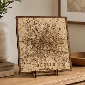 Peut inclure: Une décoration murale carrée en bois présentant une carte gravée au laser de Berlin. La carte en bois clair est encadrée d'une bordure marron foncé et comprend le texte BERLIN avec une petite silhouette d'horizon en bas, posée sur un support.