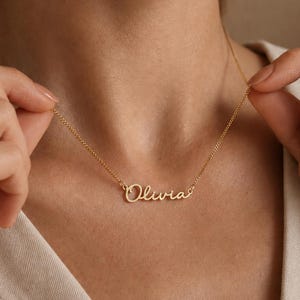 Puede incluir: Un collar de tono dorado que presenta una cadena delicada y un colgante central que muestra el nombre Olivia en una fuente cursiva. El metal tiene un acabado pulido y la placa con el nombre está integrada directamente en los eslabones.