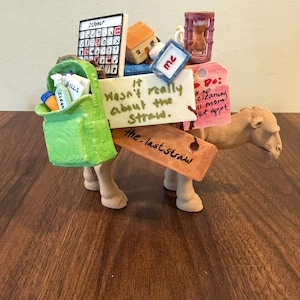 Puede incluir: Una figura de camello color canela que lleva una mochila verde llena de artículos de limpieza, un calendario marcado como octubre, una casa pequeña, un reloj de arena y etiquetas con texto que incluye It wasn't really about the Straw y the last straw.