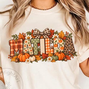 May include: A cream-colored t-shirt featuring a graphic design with the name MATILDA in block letters. Each letter has a unique pattern including plaid, floral, polka dot, and leopard print. The text is surrounded by pumpkins, leaves, and a bow.