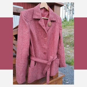 May include: A textured red blazer featuring a notched collar, three front buttons, and a matching tie belt at the waist. The garment includes two front patch pockets and a decorative circular brooch pinned to the left lapel, with a solid red lining.