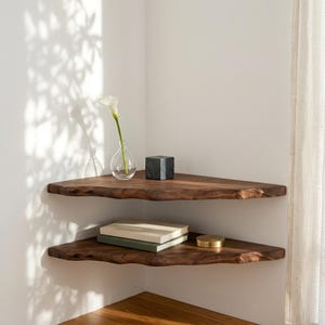 May include: Two dark brown wooden corner shelves with natural live edges mounted on a white wall. The top shelf holds a clear glass vase with a single white flower and a small black cube. The bottom shelf holds two stacked books and a gold disc.