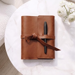 Puede incluir: Un cuaderno rectangular de color marrón con acabado liso, asegurado por una correa a juego atada con un nudo. Un bolígrafo negro mate está insertado verticalmente en una ranura en la cubierta frontal, apoyado sobre la correa anudada.