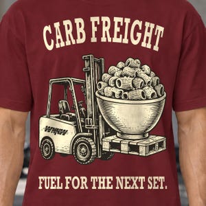 以下が含まれることがあります： マルーンカラーのTシャツで、パスタが入った大きなボウルを運ぶフォークリフトのグラフィックが描かれています。フォークリフトの上には「CARB FREIGHT」、下には「FUEL FOR THE NEXT SET.」という文字がクリーム色のフォントでプリントされています。