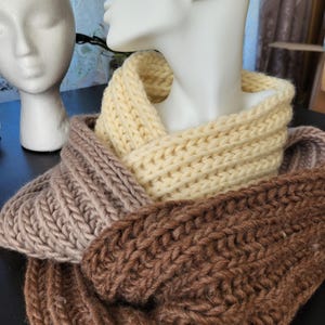 Puede incluir: Una bufanda infinita de punto grueso con tres secciones de colores distintos en crema, beige claro y marrón oscuro. La bufanda está confeccionada con una textura de hilo acanalado grueso y drapeada en capas superpuestas para crear una forma circular.