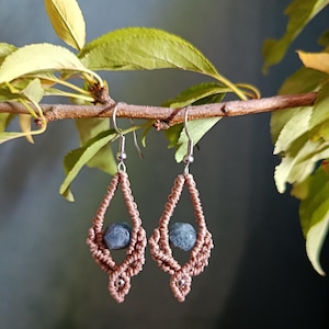 Peut inclure: Une paire de boucles d'oreilles pendantes en cordon macramé marron clair noué en forme de losange. Chaque boucle contient une perle ronde en pierre bleu-gris marbrée au centre et une petite perle en métal argenté à la pointe inférieure.
