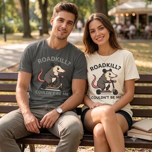 Könnte beinhalten: Ein anthrazitfarbenes T-Shirt und ein cremefarbenes T-Shirt, beide mit einer Grafik eines Opossums auf einem Roller und dem schwarzen Schriftzug ROADKILL? COULDN'T BE ME. Die T-Shirts haben kurze Ärmel und einen Rundhalsausschnitt mit Cartoon-Motiv.