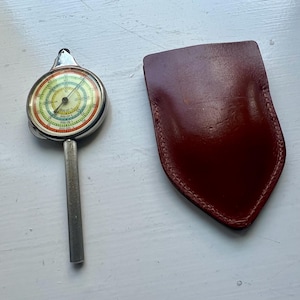 Op de afbeelding: Een cirkelvormige mechanische meter met een zilverkleurige metalen handgreep en een veelkleurige wijzerplaat met concentrische ringen en numerieke markeringen, vergezeld van een gladde, donkerbruine lederen beschermhoes met een puntige onderkant.