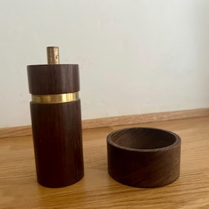 May include: A cylindrical dark brown wooden pepper mill featuring a horizontal brass band around the center and a small brass knob at the top, accompanied by a separate, matching low-profile circular wooden bowl with a visible grain pattern.