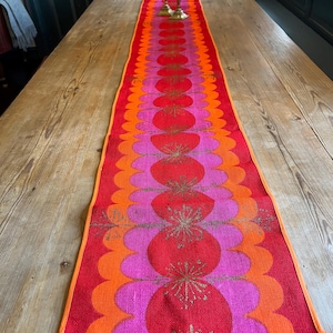 Può includere: Un lungo runner da tavola rettangolare caratterizzato da un vivace motivo di cerchi sovrapposti nelle tonalità del rosso brillante, rosa acceso e arancione. Il design include motivi dorati ripetuti a forma di stella o fiocco di neve.